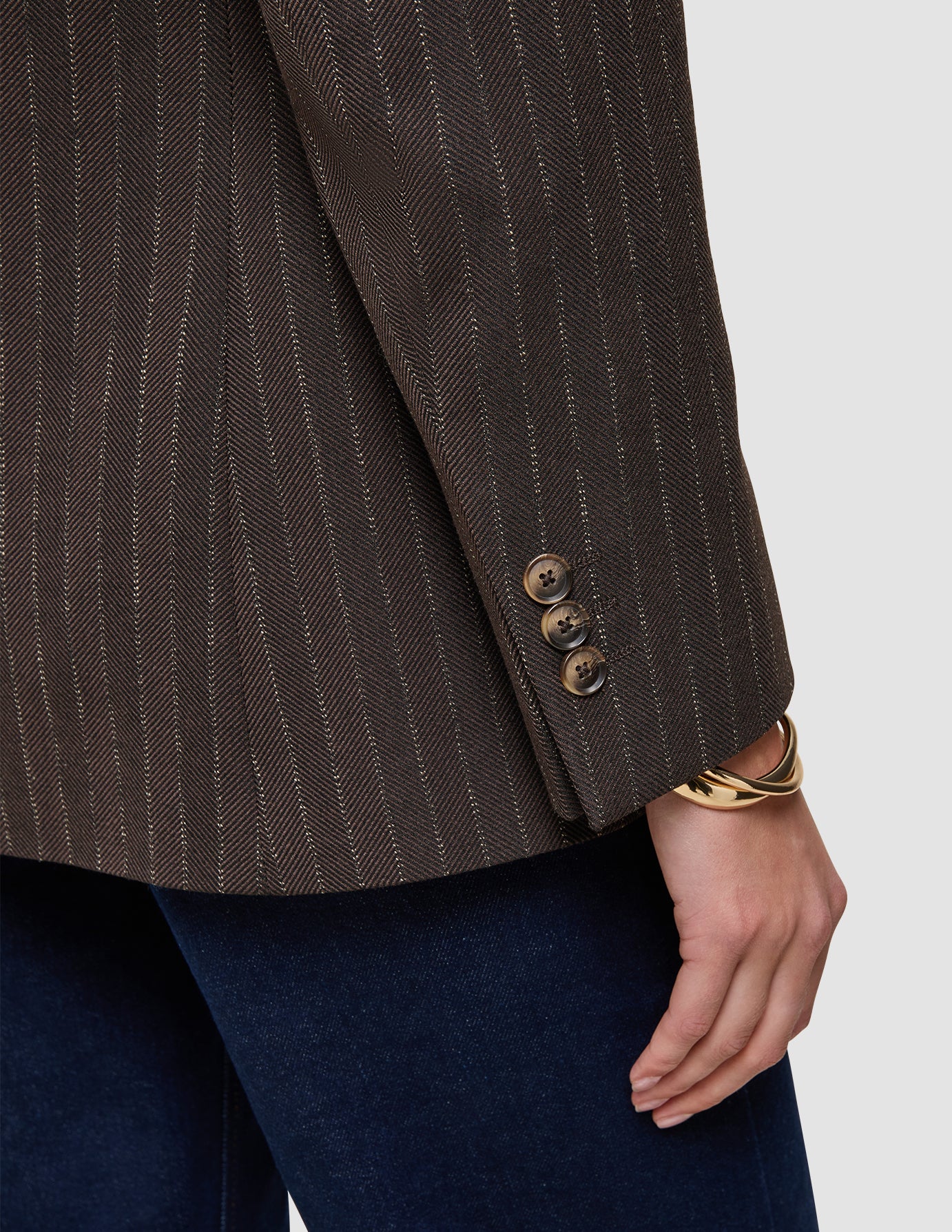 Nahaufnahme eines Women Serene-Blazers in der Passform Regular in der Farbe Walnut mit Fischgrätenmuster am Ärmel, brauner Fischgrätenstruktur in Wolloptik, drei Knöpfen an den Manschetten und einem goldenen Armreif am Handgelenk.